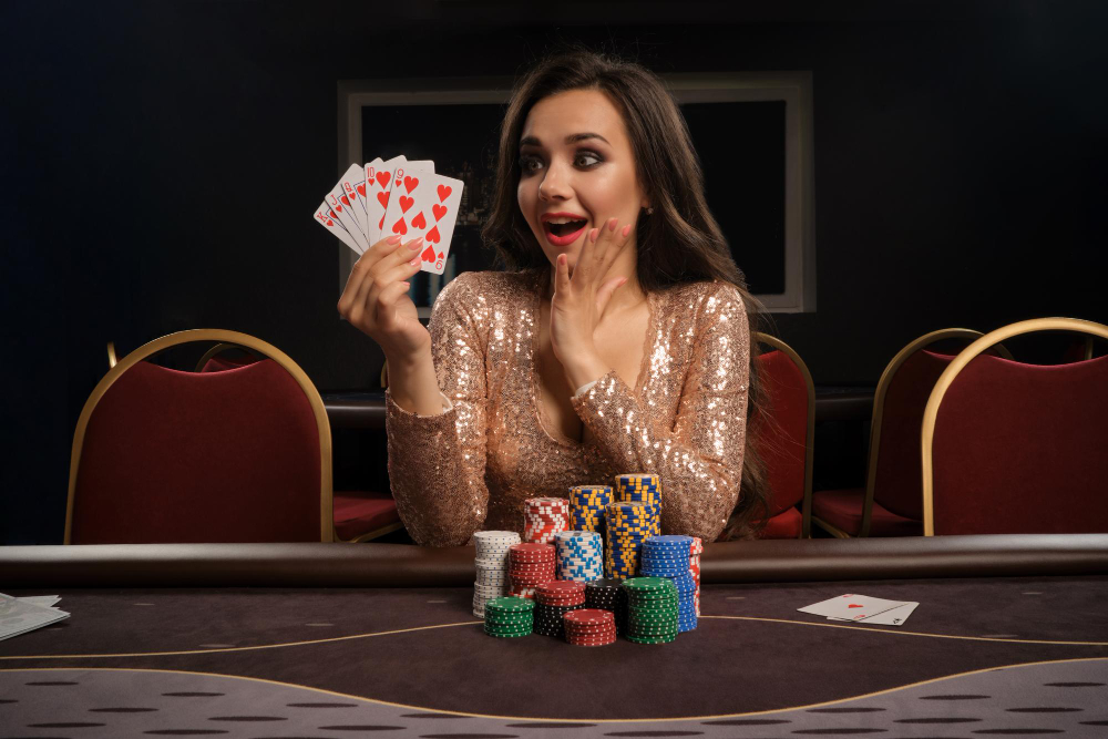 Happy brunette girl in a brilliant gold dress is playing poker sitting at the table in casino. She is making bets waiting for a big win and showing her cards. Gambling for money. Games of fortune.
