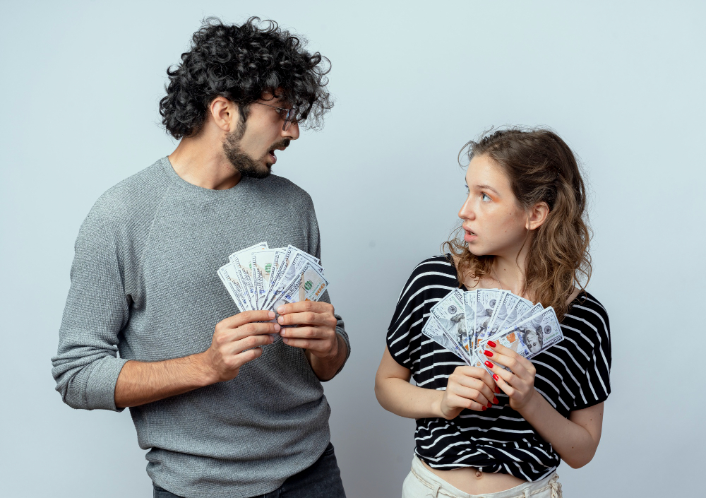 Young beautiful couple man and women holding cash looking at each other surprised standing over white wall
