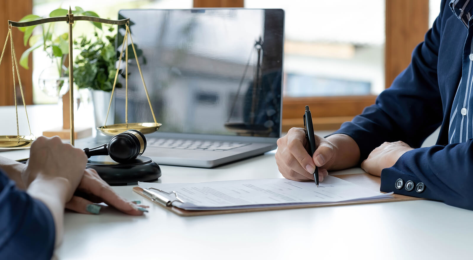 Person on the right writing on a paper and person on the left is sitting next to a judges mallet and the scale of justice with their hands crossed 