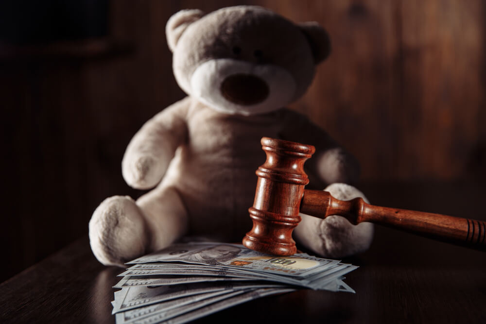 A stack of money with a judges mallet on top of it and a teddy bear behind with dark lighting