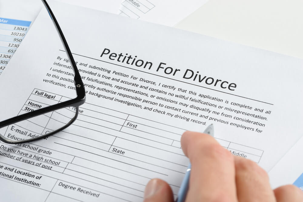 Petition for divorce paper with glasses laying on top of it and someones hand with a pen about to fill out the paper