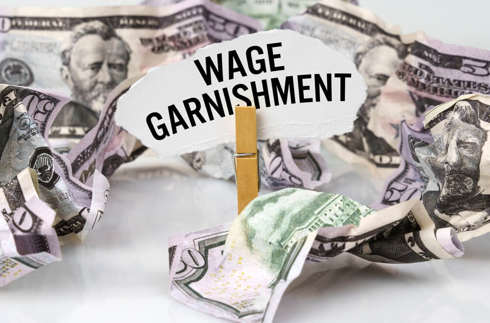 The word wage garnishment on a cut out piece of paper help up on a close-pin with money crumbled around it 