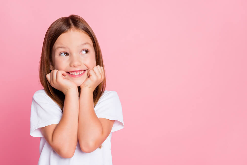 Little girl standing in front of a pink background with her hands resting on her chin and a smile on her face