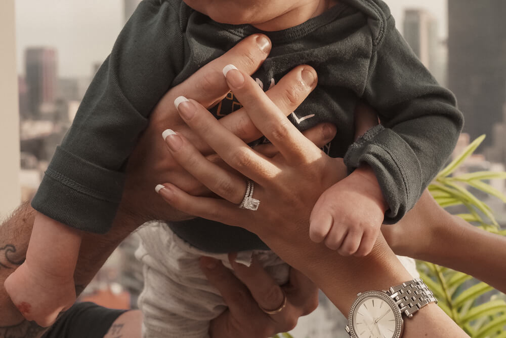 Close up of a child and the mother and fathers hands are holding the baby up, mother has a wedding ring on 