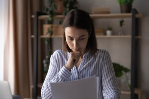 Women sitting at a desk looking over some papers, one hand is holding the papers and the other hand has her chin resting on it 