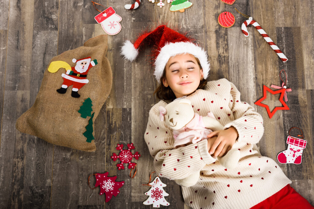 Girl in a Santa cap laying on the floor with her eyes closed holding a stuffed animal