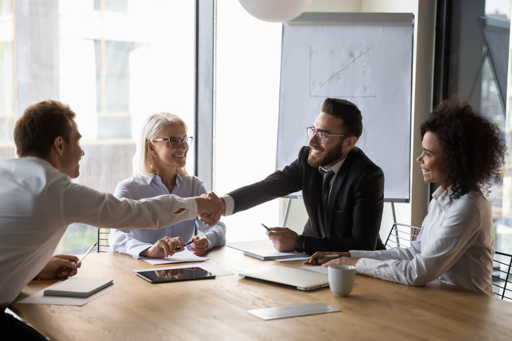 Four people, two women and two men sitting at a table and the two men are shacking hands