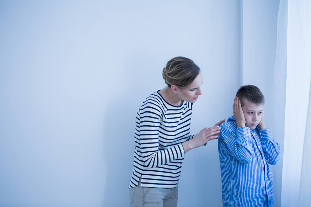 Mother behind her son talking to him and the boy has his ear covered with his hands 