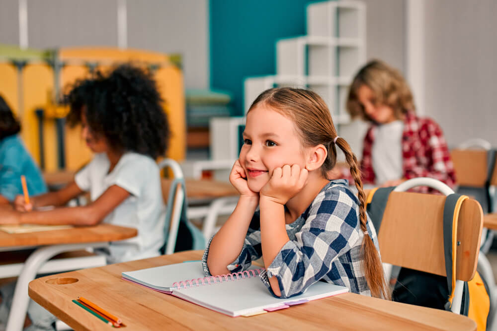 Little girl sitting at school desk with her notebook open and her elbows in the desk and head resting on her hands with 2 kids working on the background