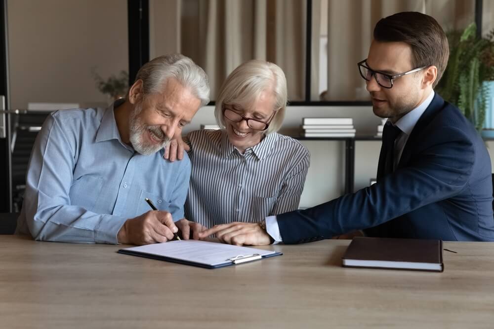 An older couple sitting at a desk with a clipboard in front of them and a man next to the couple showing them where ti sign 