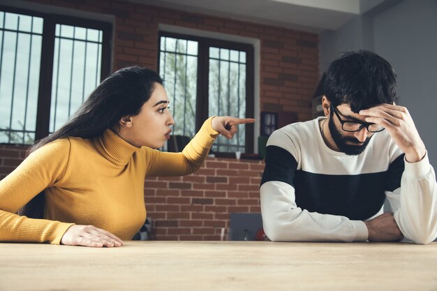 In a room with brick walls and windows, a woman in a yellow sweater points at a man with glasses who appears upset, resting his head in his hand. It seems she's highlighting one of the Texas divorce grounds, adding tension to the scene.