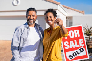 Man and women standing in front of a house and a sign that says for sale with a sold sticker over it and the women is holding up the keys to the house