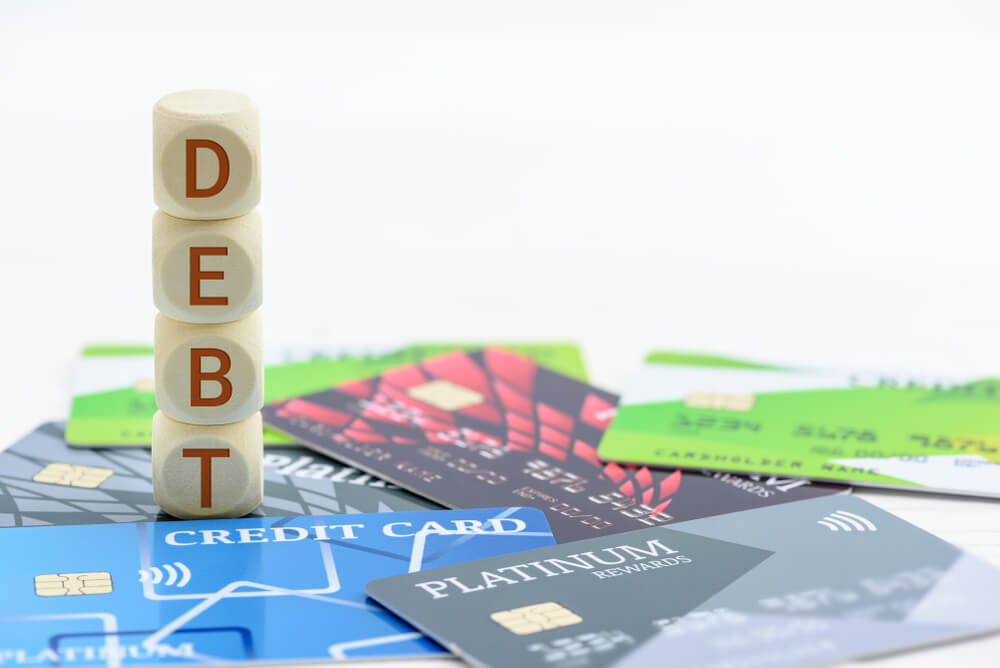 Four blocks stacked on top of each other spelling out the word debt from the top block to the bottom block and the blocks are around many credit cards spread out on the table