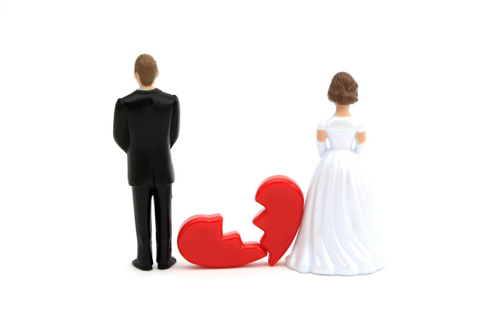 A groom and bride wedding cake toppers standing next to a red heart that is cut in have jaggedly representing a broken heart