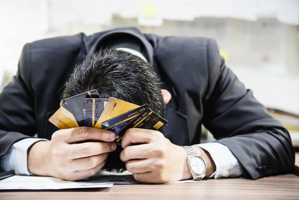 Businessman sitting at a desk resting his head on the desk while holding up may fanned out credit cards