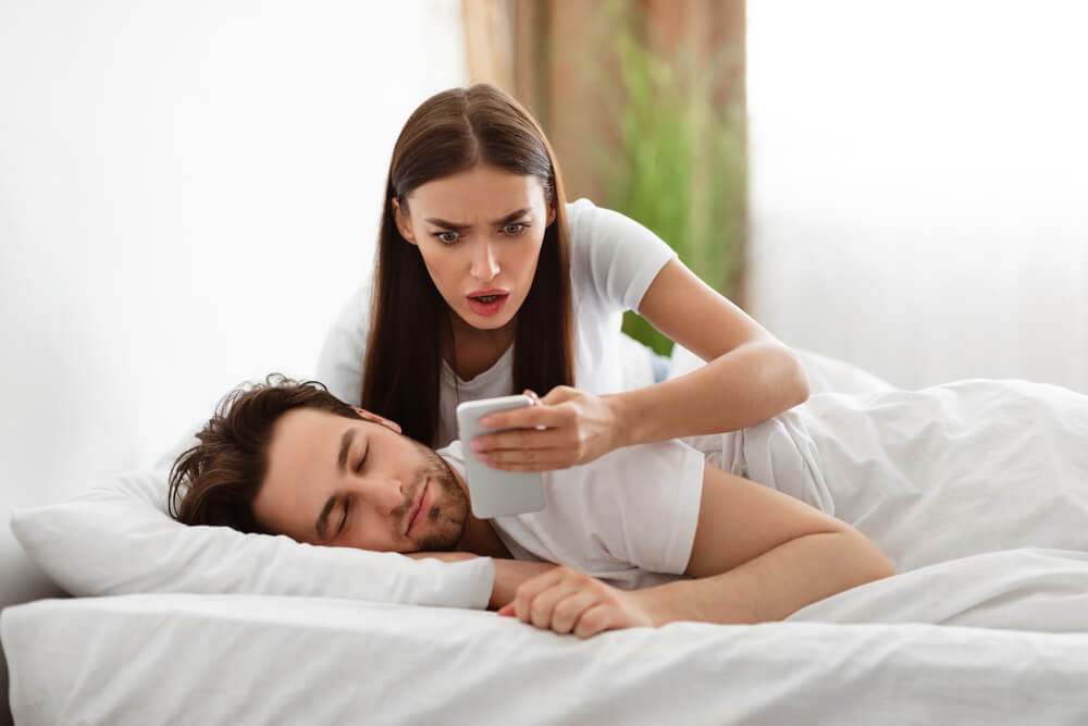 A man is sleeping in bed and a woman comes up behind him and looks at his phone, the woman seems chocked and angry 