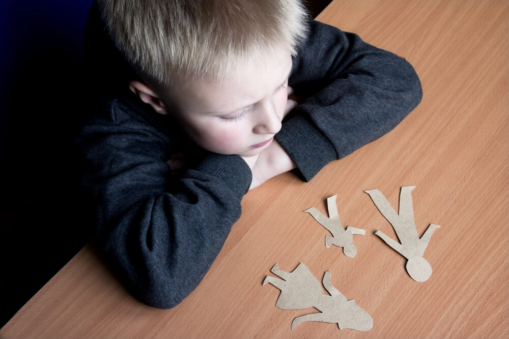 A little boy laying is arms on a wood table and his head resting on his arms looking at people cutouts of a mom, dad, and child