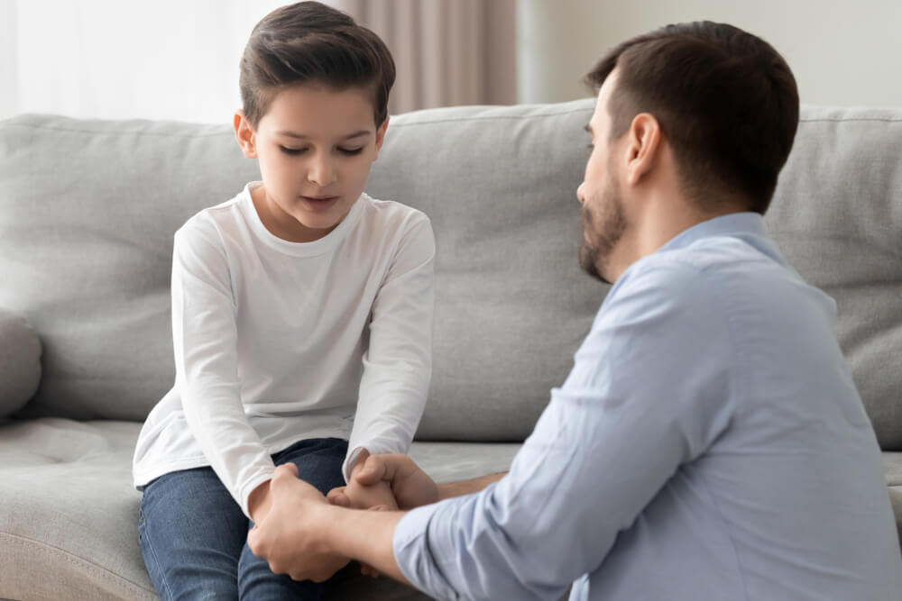 Little boy sitting on a grey couch holding his dads hands and his dad is kneeling on the floor text to him
