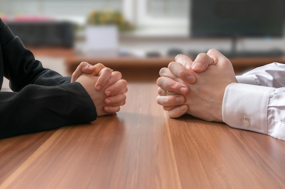 A close up of two people sitting at a wooden table and both of their hands are clasped