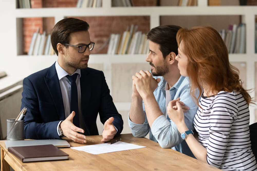 Husband and wife sitting at a wooden table talking with an attorney with a paper, a book, and pens sitting on the desk