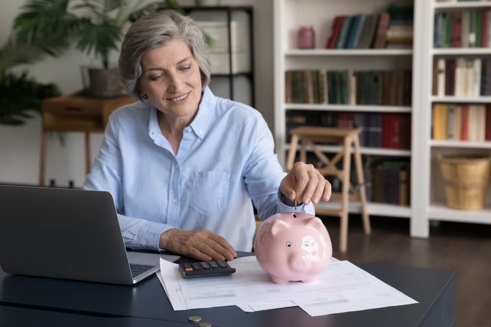 Women adding money to a piggybank that is on top of papers and next to her open computer and a calculator 