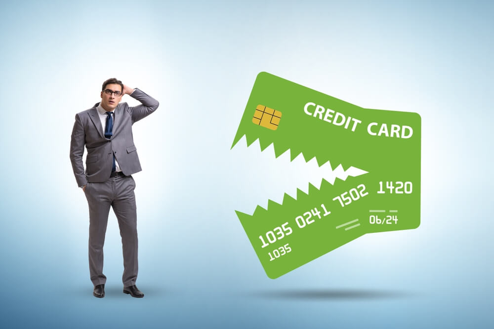A businessman standing to the left with one hand in his pocket and the other on his hid and a big credit card cut in half jaggedly looking like the card is going to eat the man