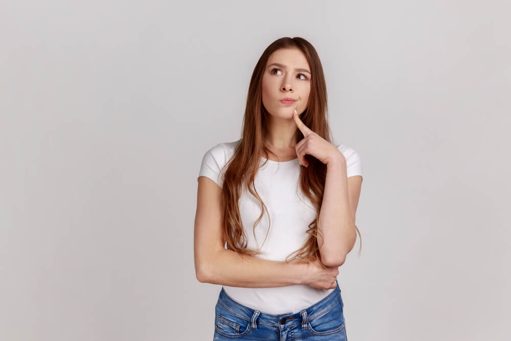 A woman standing against a white background looking up and to the side like she is making a decision 
