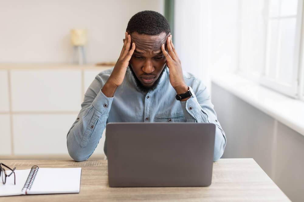 A man sitting at his desk looking at his open laptop and his elbows are resting on the desk and his hands are on his temples 