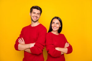 A man and women standing against a yellow wall with their backs together and their arms crossed 