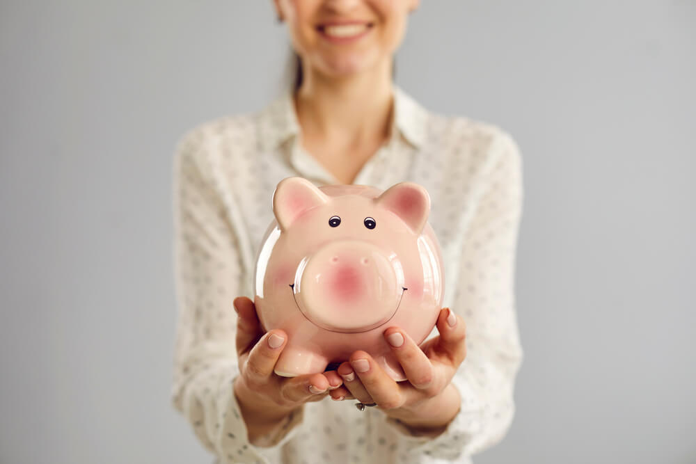 A woman holding a pink piggy bank with both of her hands. She is holding the piggy bank away from her body offering it. 