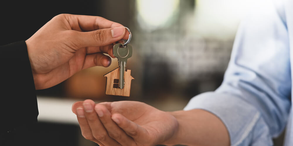 A close up of hands and the hand on the left is holding a key with a house key chain and is placing the keys in the person to the rights hand