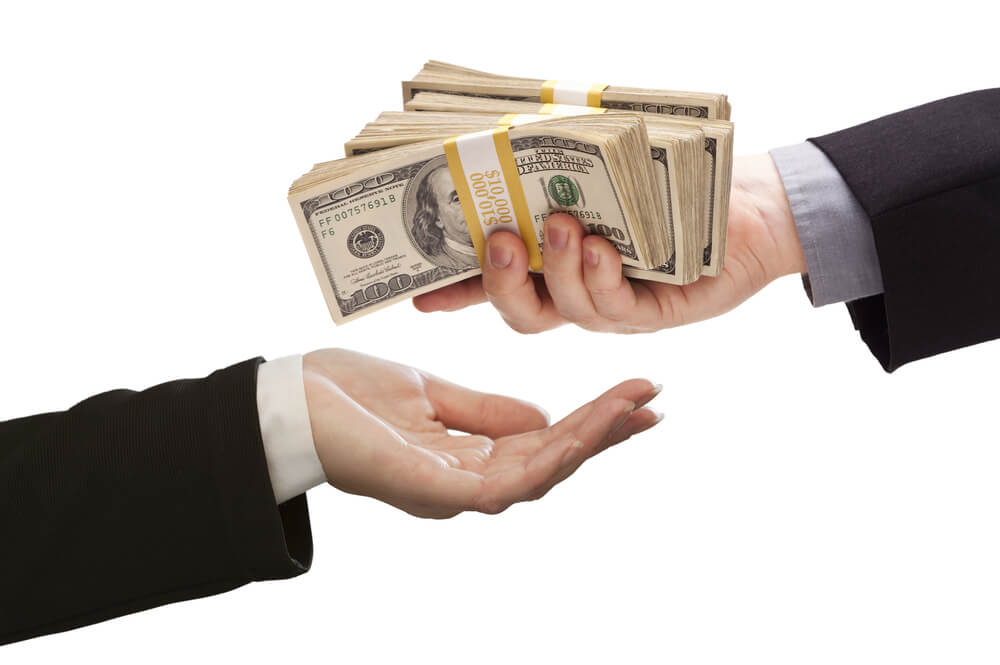 A close up of two hands, the one on the right is holding stacks of hundred dollar bills and the one on the left has their hand out waiting for the money