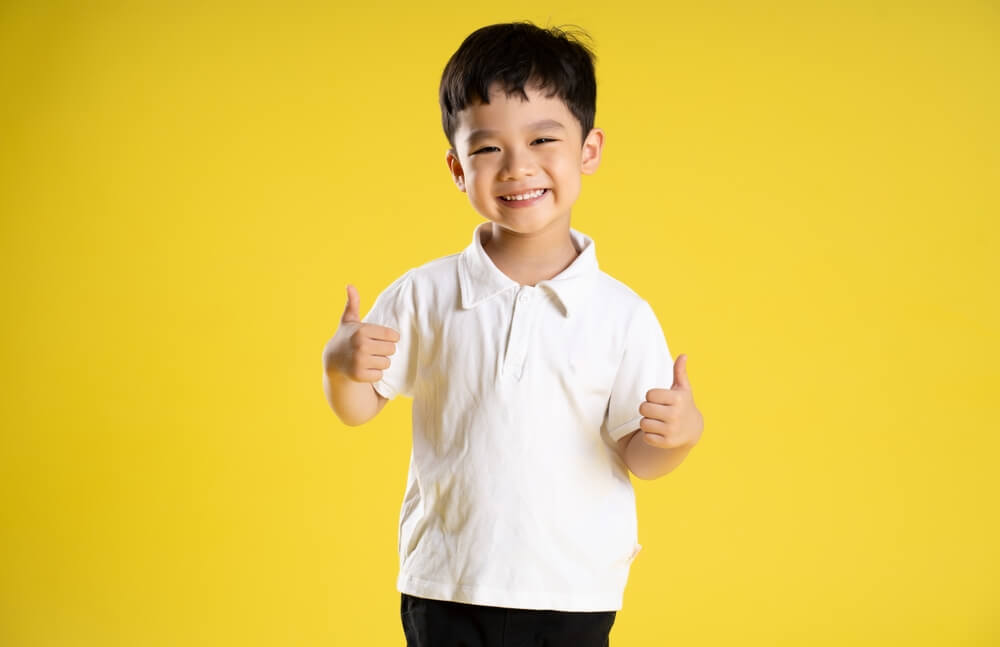 A little boy smiling with both of his thumbs up standing against a yellow background 
