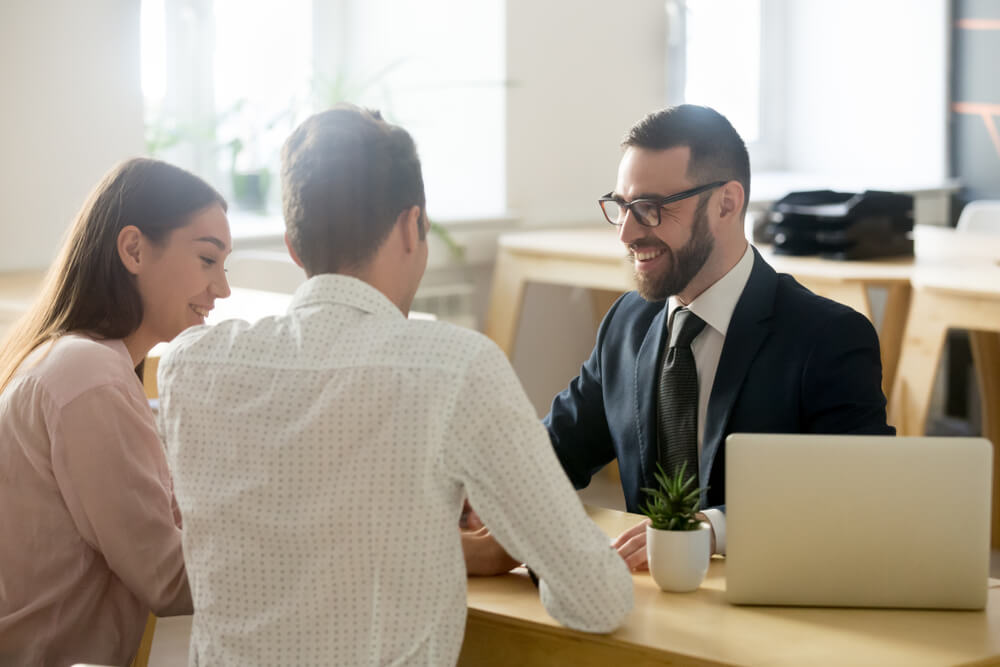 A man and woman standing on on side of a desk and on the other side is a businessman with his laptop open talking to the couple