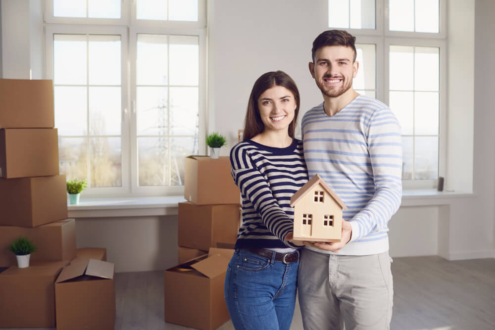 Man and woman standing next to each other both holding a wooden toy house with many brown boxes behind them 