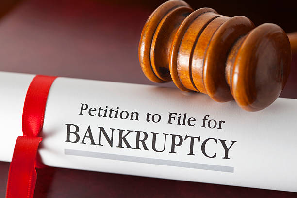 A rolled document labeled "Petition to File for Bankruptcy," hinting at questions like how much debt you need to file Chapter 7, is tied with a red ribbon, resting beside a wooden gavel.