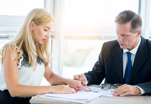 A woman and a man in business attire are seated at a table, reviewing what estate planning documents they need, with pens in hand and glasses resting on the table.