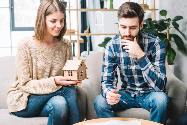 A woman holds a small house model while sitting beside a man with various papers spread on the table in front of them.