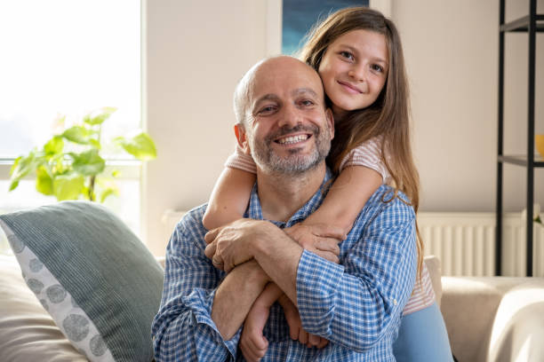 A smiling man in a blue checked shirt sits on a couch, while a joyful girl hugs him from behind in the brightly lit room, showcasing their strong bond—a bond essential for smooth co-parenting.