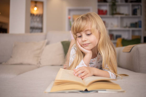 A young girl with long blonde hair is lying on a beige sofa, immersed in a book, her head resting on her hand as she escapes into another world. Bookshelves stand proudly in the background, whispering tales of harmony and the complexities of co-parenting.
