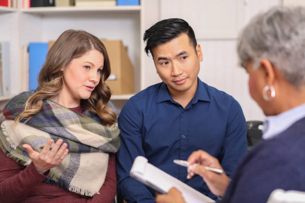 A couple sits across from a counselor, engaged in a discussion about why divorce is so expensive. The woman gestures with her hand while the man listens attentively. The counselor takes notes on a clipboard, offering guidance through their financial considerations.