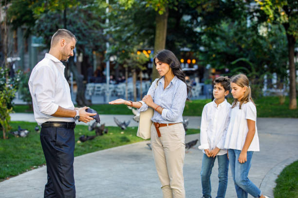 A man hands a wallet to a woman outdoors, with two children standing nearby, perhaps discussing how to get spousal support in Texas. They are in a park, surrounded by grass and trees, on a sunny day.