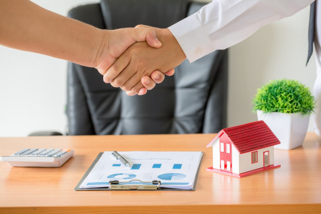 Two people shaking hands over a desk, where a trust-based estate plan takes shape amidst a document, model house, calculator, and potted plant.