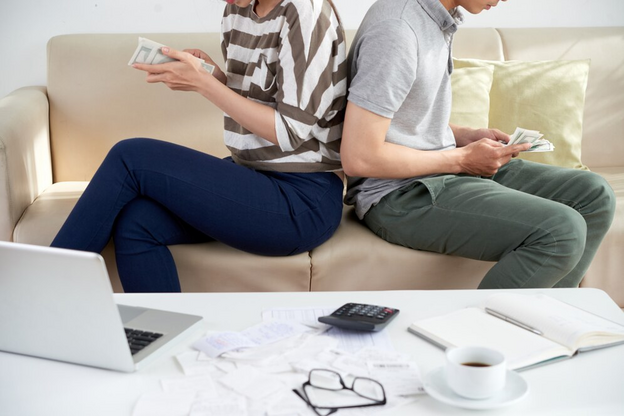 A man and woman sitting on a couch holding money and a laptop.