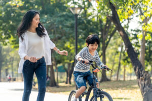 Little boy riding a bike in a park and his mother is behind him watching to make sure he does not fall