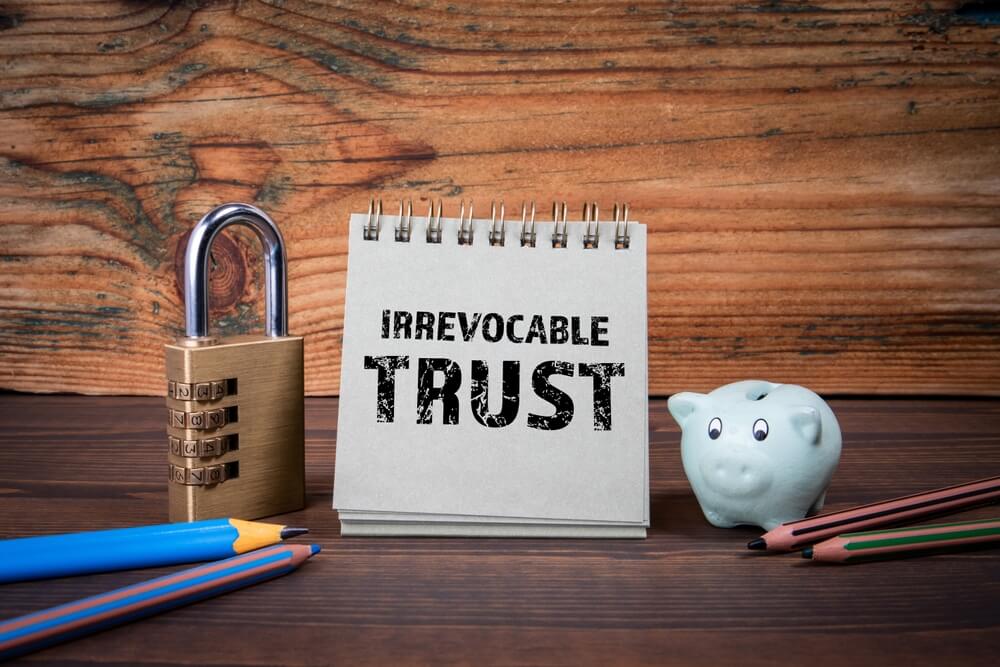 A cardboard notepad that says irrevocable trust sitting next to a closed lock and two pencils on one side and a piggy bank and two more pencils on the other side 