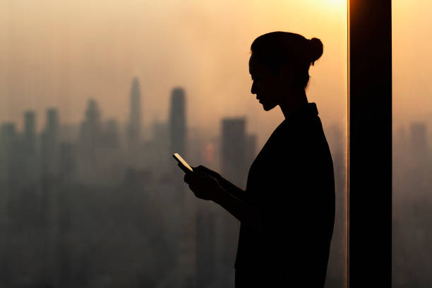 Silhouette of young woman using smartphone next to window with cityscape, Shenzhen, China