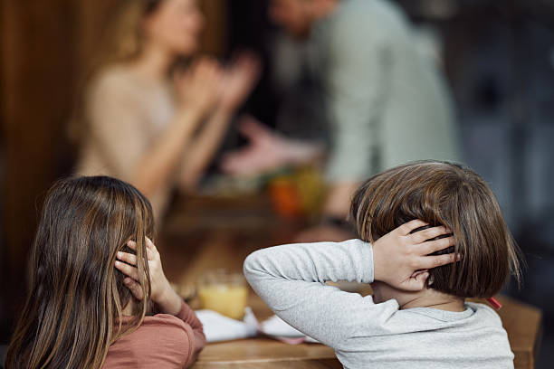Rear view of small siblings covering their ears while refusing to listen their parents arguing in dining room.