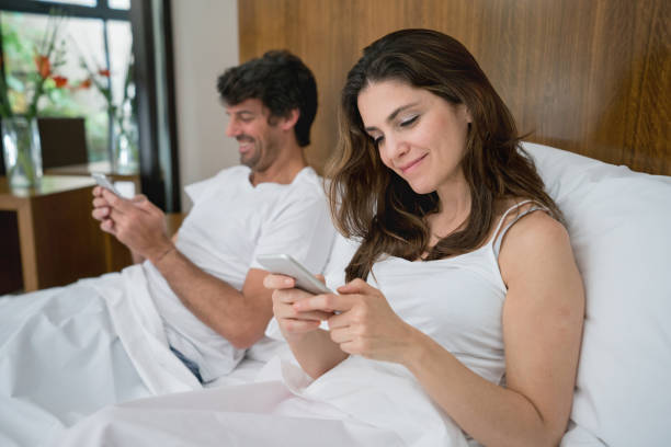 Couple sitting in bed chatting on their smartphones looking very happy and smiling, How Social Media May affect your Divorce case