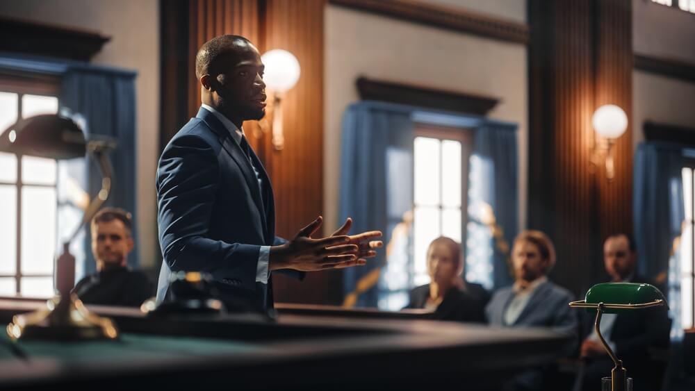Male lawyer standing in a court room talking with people in the background sitting down 